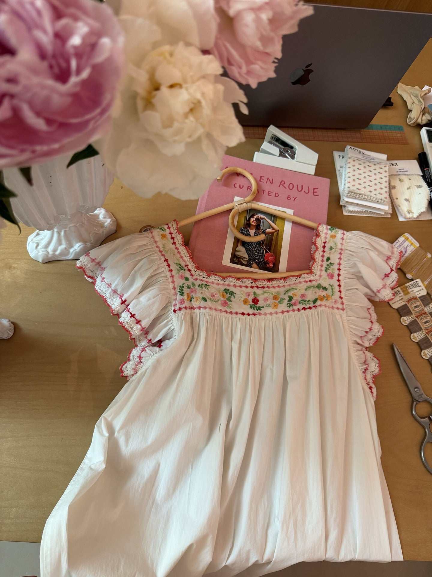 A white cotton smocked blouse with pink and green floral embroidery around the neckline, displayed on a wooden hanger on a desk with flowers, a laptop, and stationery in the background.