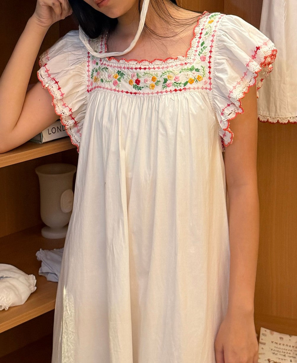 A woman wearing a white short-sleeve dress with colorful floral embroidery around the neckline and ruffled trim on the sleeves, standing in a wooden interior.