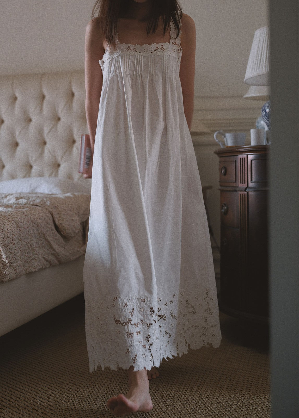 A woman wearing a white cotton nightgown with lace trim at the neckline and hem, standing barefoot in a bedroom next to a bed and a wooden nightstand.
