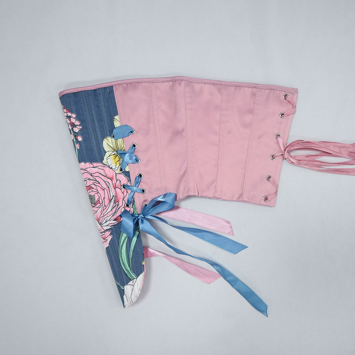 A handmade corset-style top laid flat on a light gray background. The left side features a floral print with pink, blue, and green tones, while the right side is solid pink with metal eyelets and pink ties.