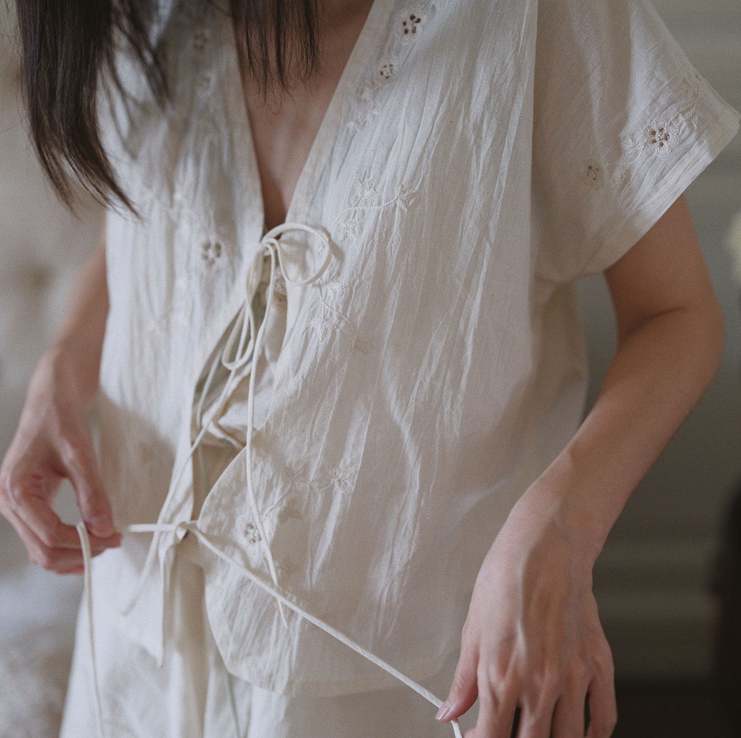 A close-up of a person wearing a loose, short-sleeve cream-colored blouse with delicate embroidery and a front tie closure, shown in a softly lit indoor setting.
