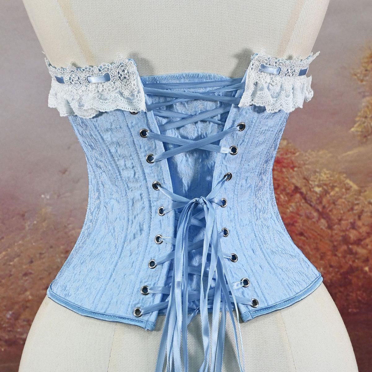 A light blue lace-up corset displayed on a cream mannequin torso, featuring textured fabric, decorative white lace shoulder straps with blue ribbons, and silver-tone eyelets.