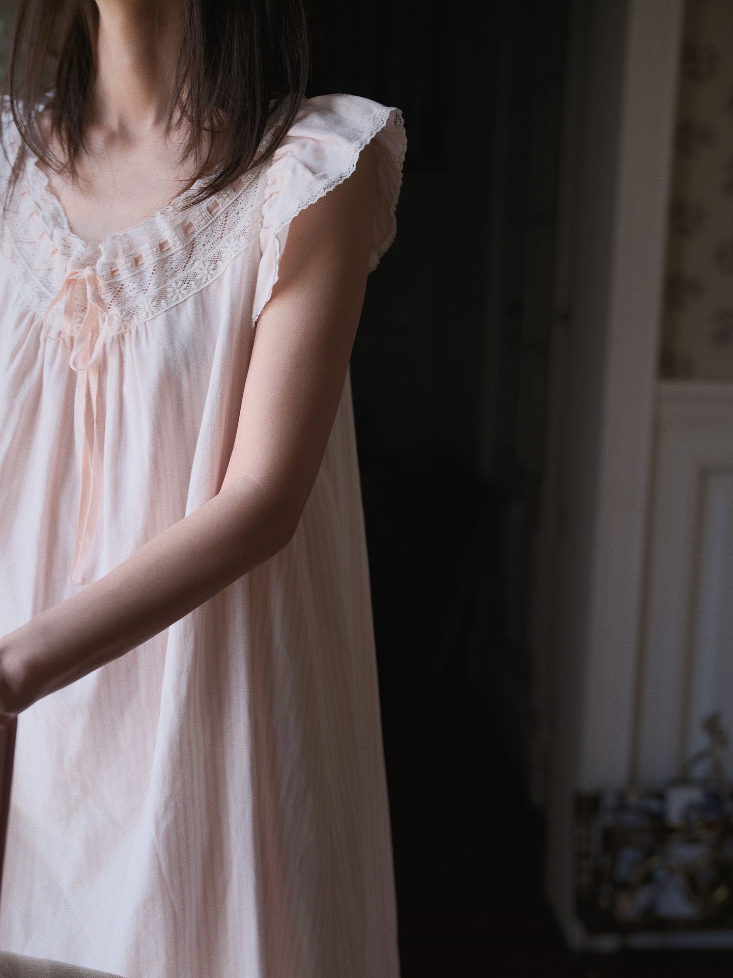 A woman wearing a pale blush pink nightgown with delicate lace trim at the neckline, shown in a softly lit indoor setting.