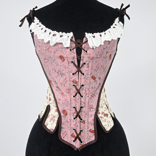 A pink floral corset bustier displayed on a black dress form against a light gray background. The corset features a pink base with a brown and blue floral pattern, white lace trim at the bust, and dark brown laces and trim.