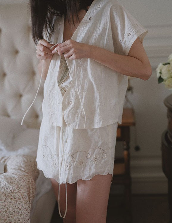 A woman wearing a cream-colored short-sleeve eyelet lace pajama set with a matching tie-front cover-up and shorts, standing in a softly lit bedroom.