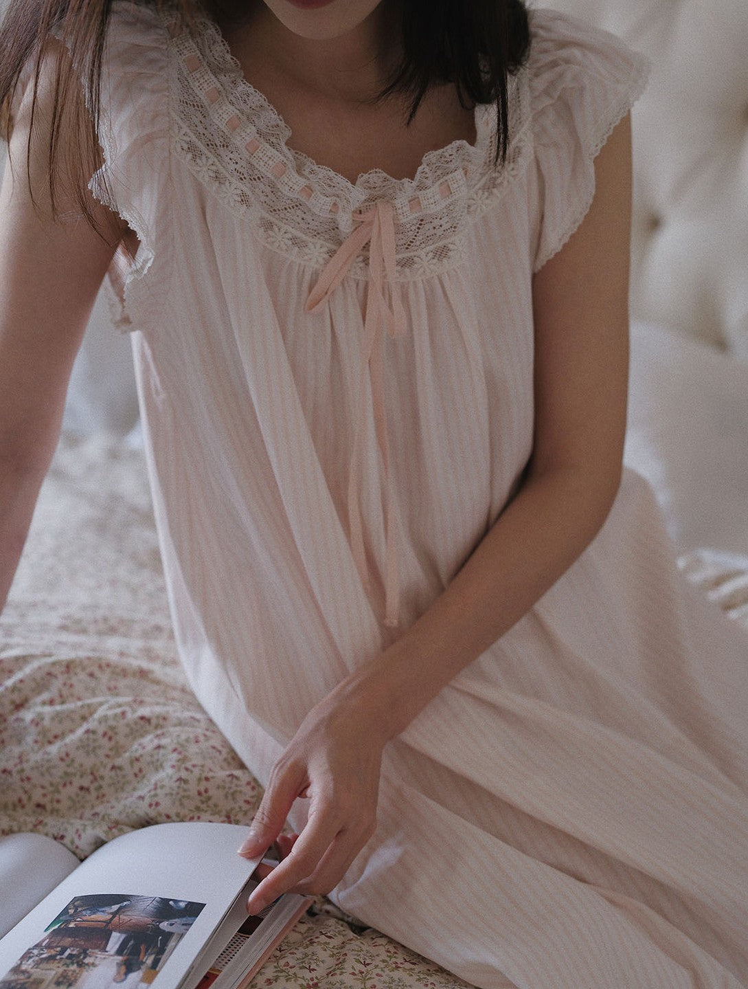 A woman wearing a pale blush pink nightgown with delicate lace trim and a tie at the neckline, sitting on a bed and holding an open book.