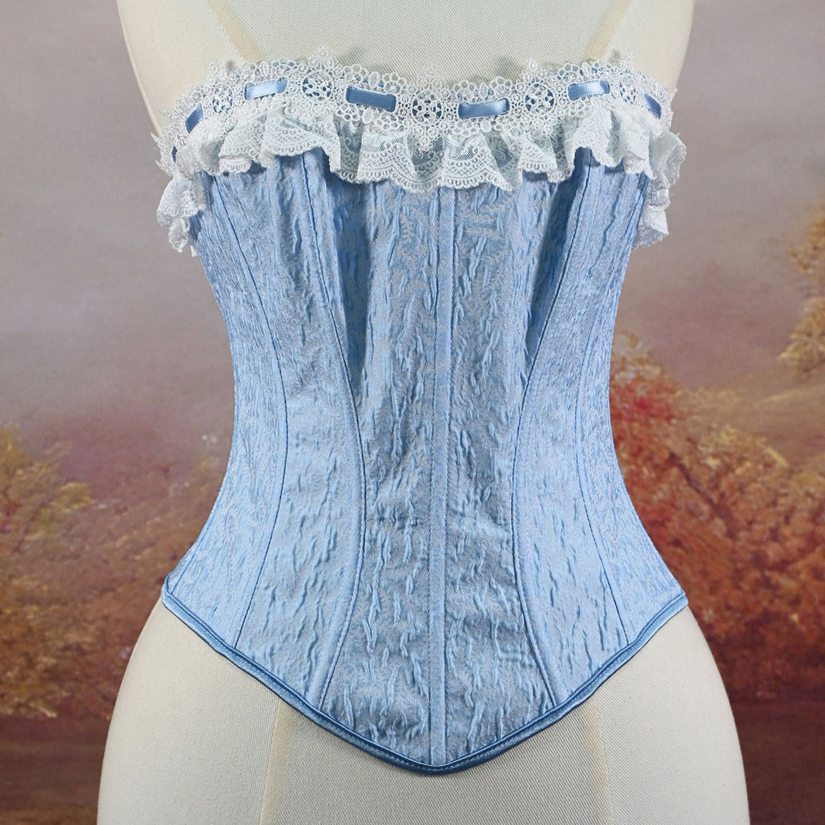 A light blue textured corset with white lace trim and ruffles, displayed on a cream-colored mannequin torso against a soft-focus autumn landscape background.