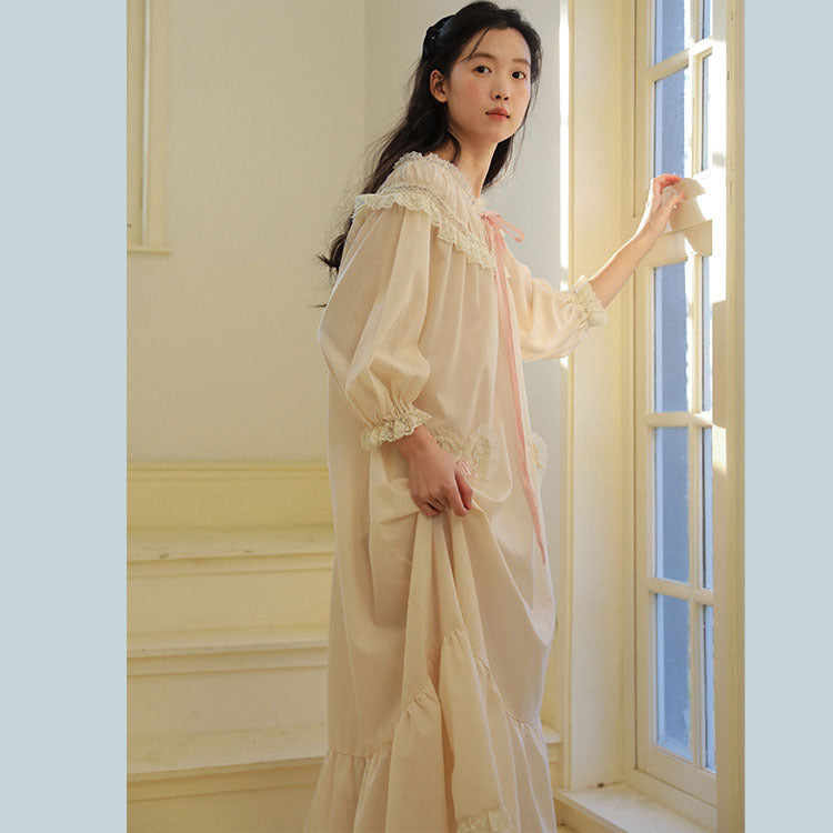 A young woman stands by a window wearing a long cream-colored nightgown with lace trim and a pink ribbon tie. The scene is set in a softly lit room with pale yellow walls and a window frame.