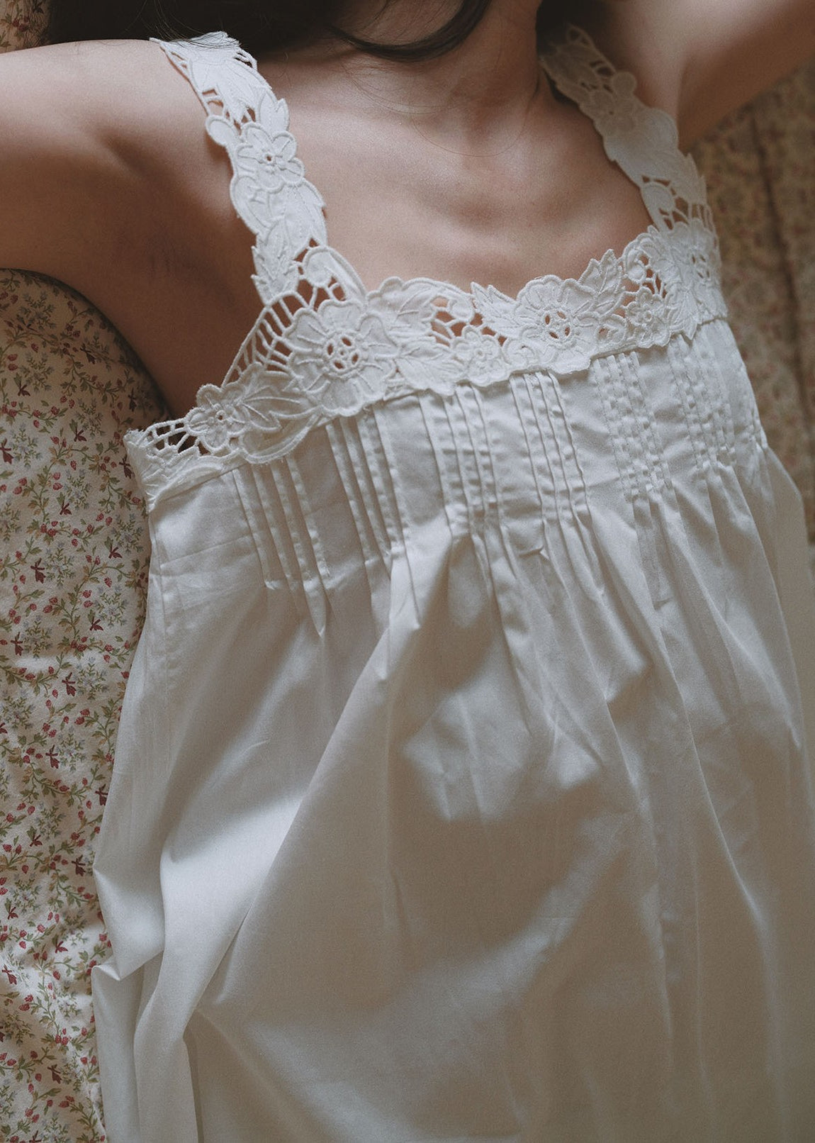 A close-up of a person wearing a white lace-trim camisole nightgown or slip, photographed indoors against a soft floral fabric background.