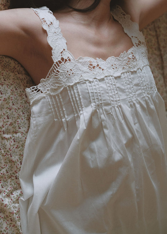 A close-up of a person wearing a white lace-trim camisole nightgown or slip, photographed indoors against a soft floral fabric background.