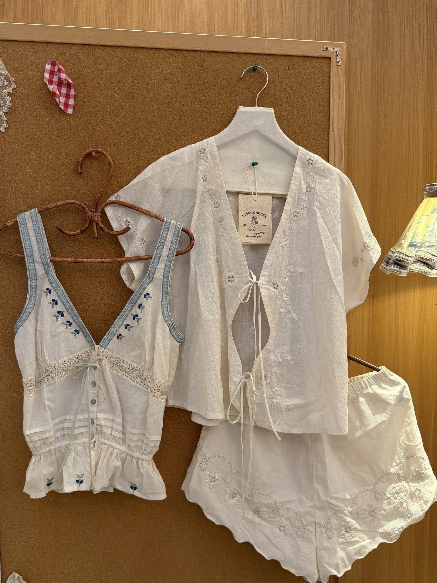 A white embroidered two-piece lingerie set with a short-sleeve lace-up cover-up top and matching ruffled shorts, displayed on hangers against a brown cork board.
