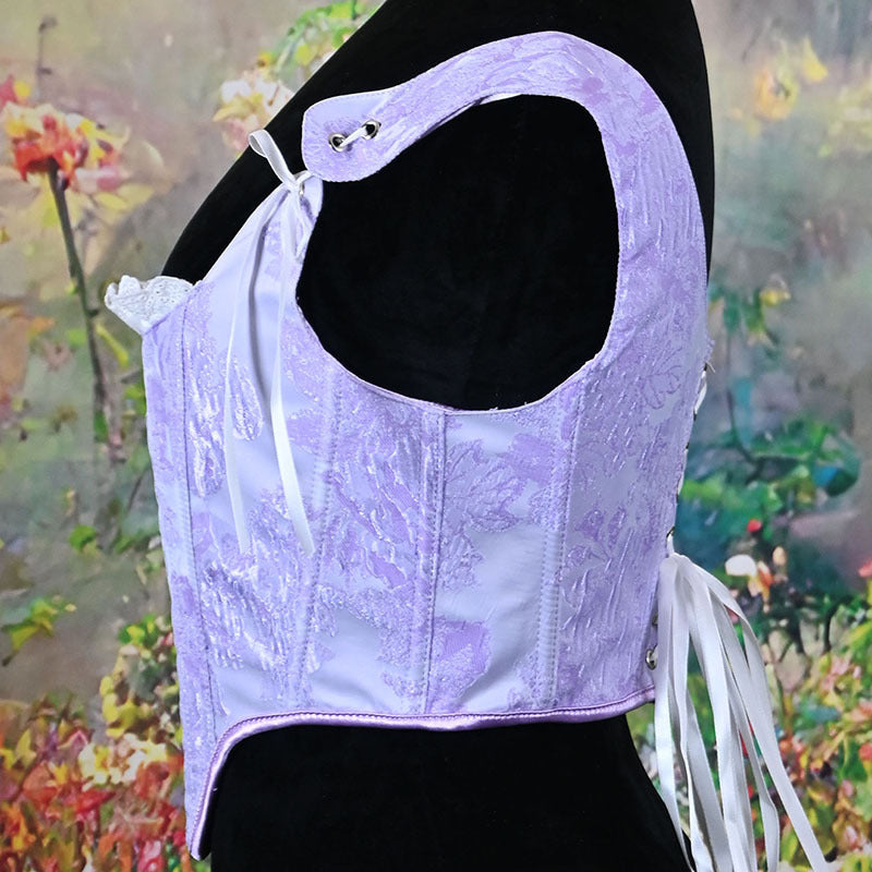 A lavender lace corset-style top displayed on a black mannequin torso against a blurred floral background. The garment has a large open back, side ties, and a fitted silhouette.