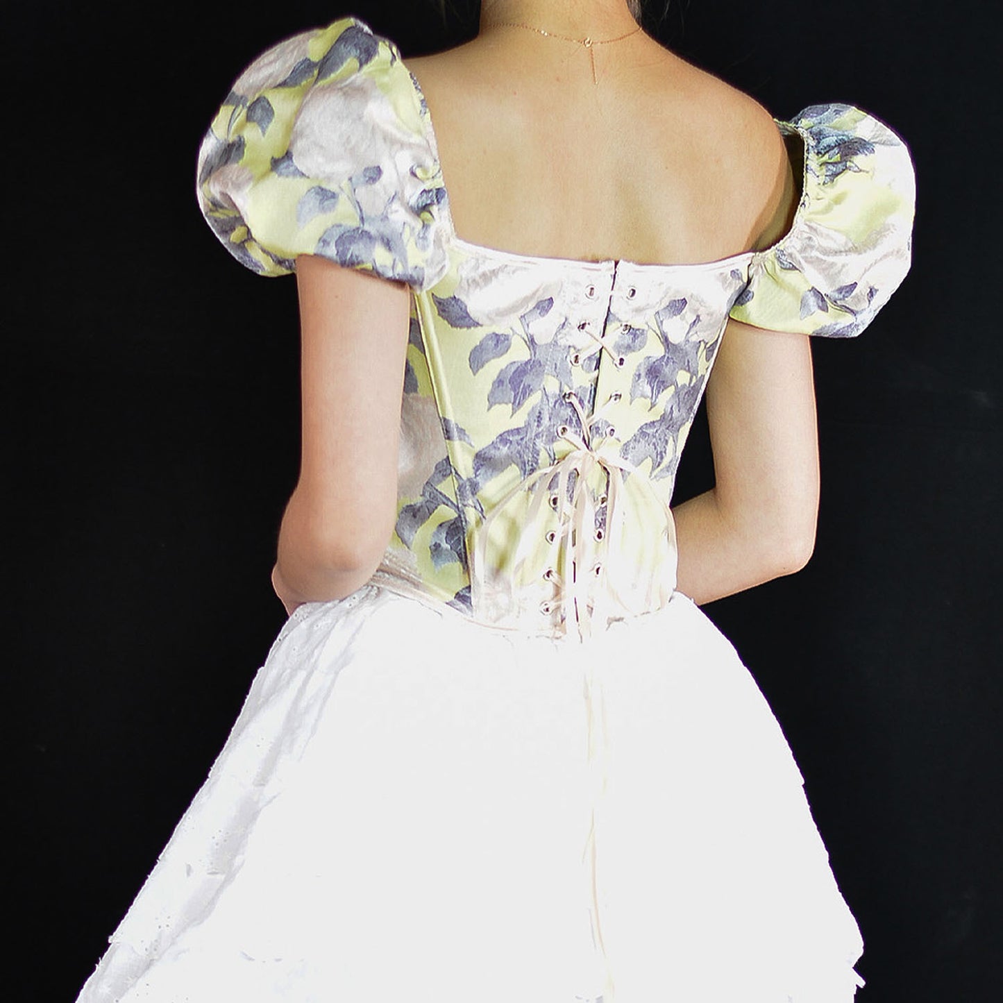 Rear view of a woman wearing a corset-style dress with puff sleeves, a pale yellow and lavender floral print bodice, and a white skirt against a black background.