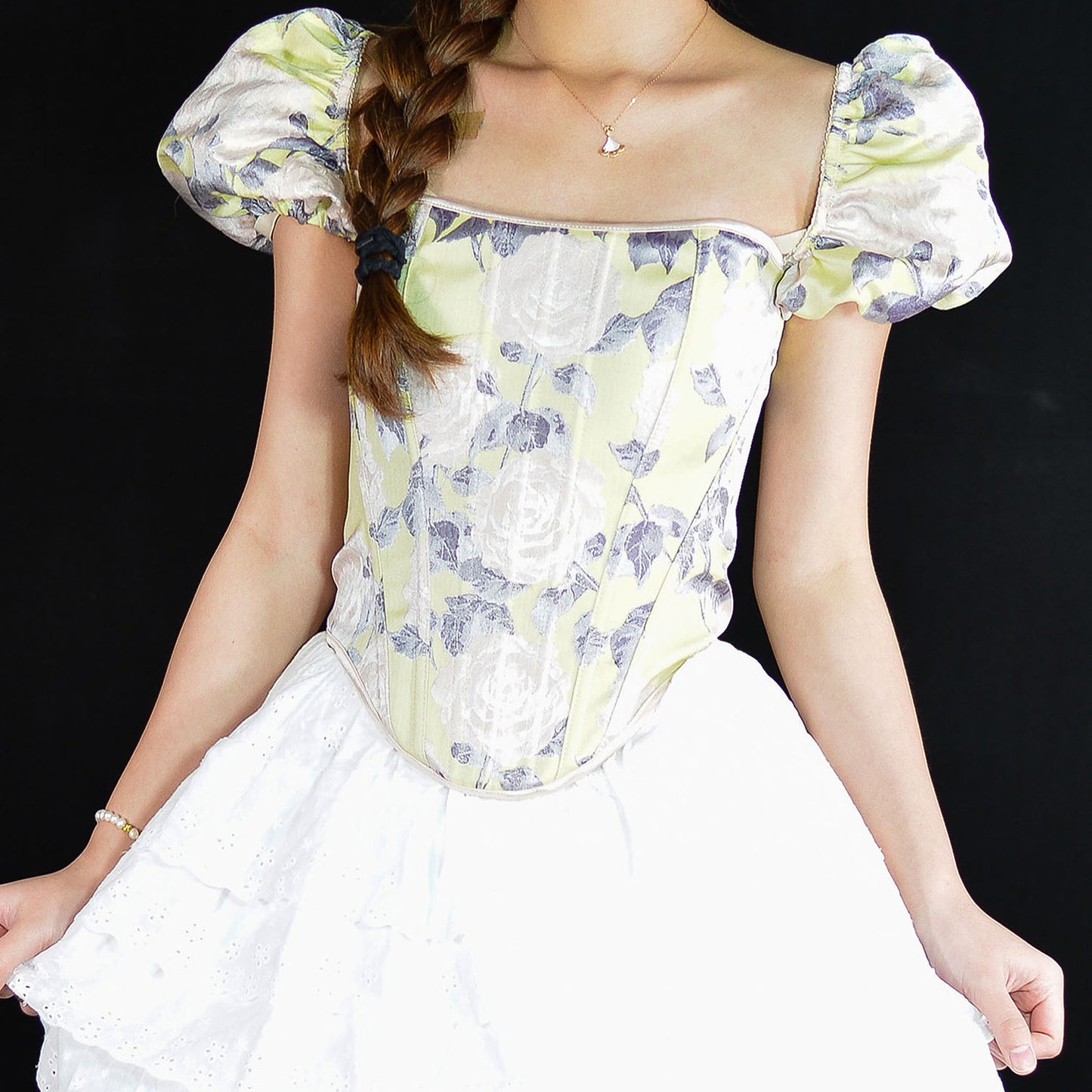 A woman wearing a white corset-style top with a pale yellow and purple floral print, paired with a white skirt, set against a solid black background.