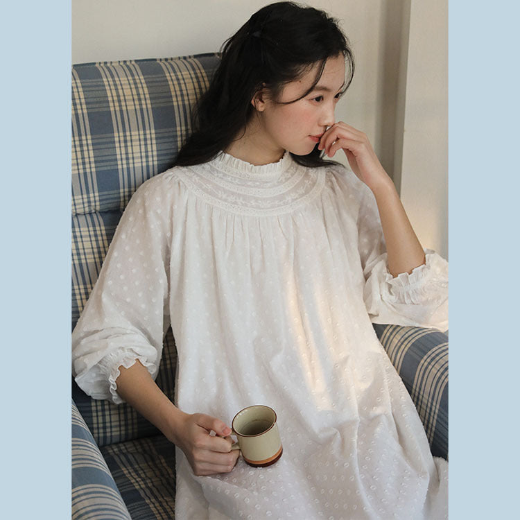 A woman sits in a blue and beige plaid armchair, wearing a white long-sleeve dress with a subtle polka dot pattern and holding a ceramic mug.