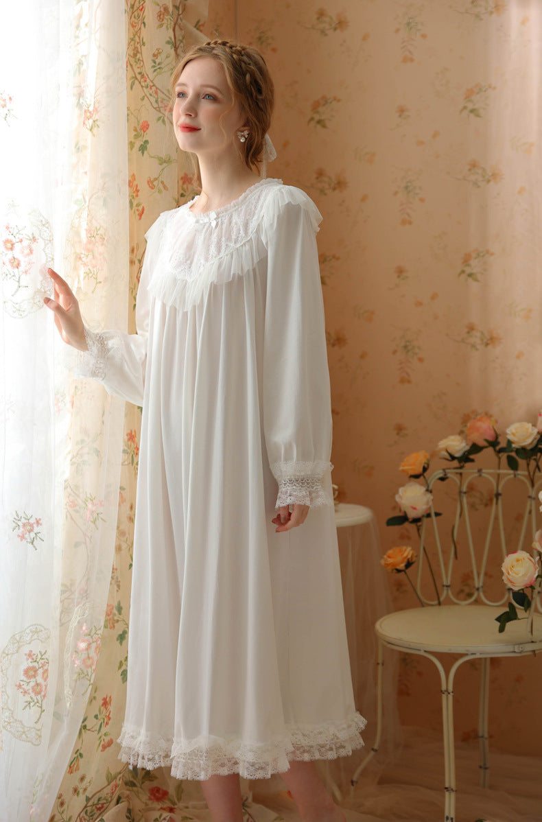 A woman wearing a long white nightgown with lace trim stands by a window in a vintage-style room with floral wallpaper and a small table with flowers.