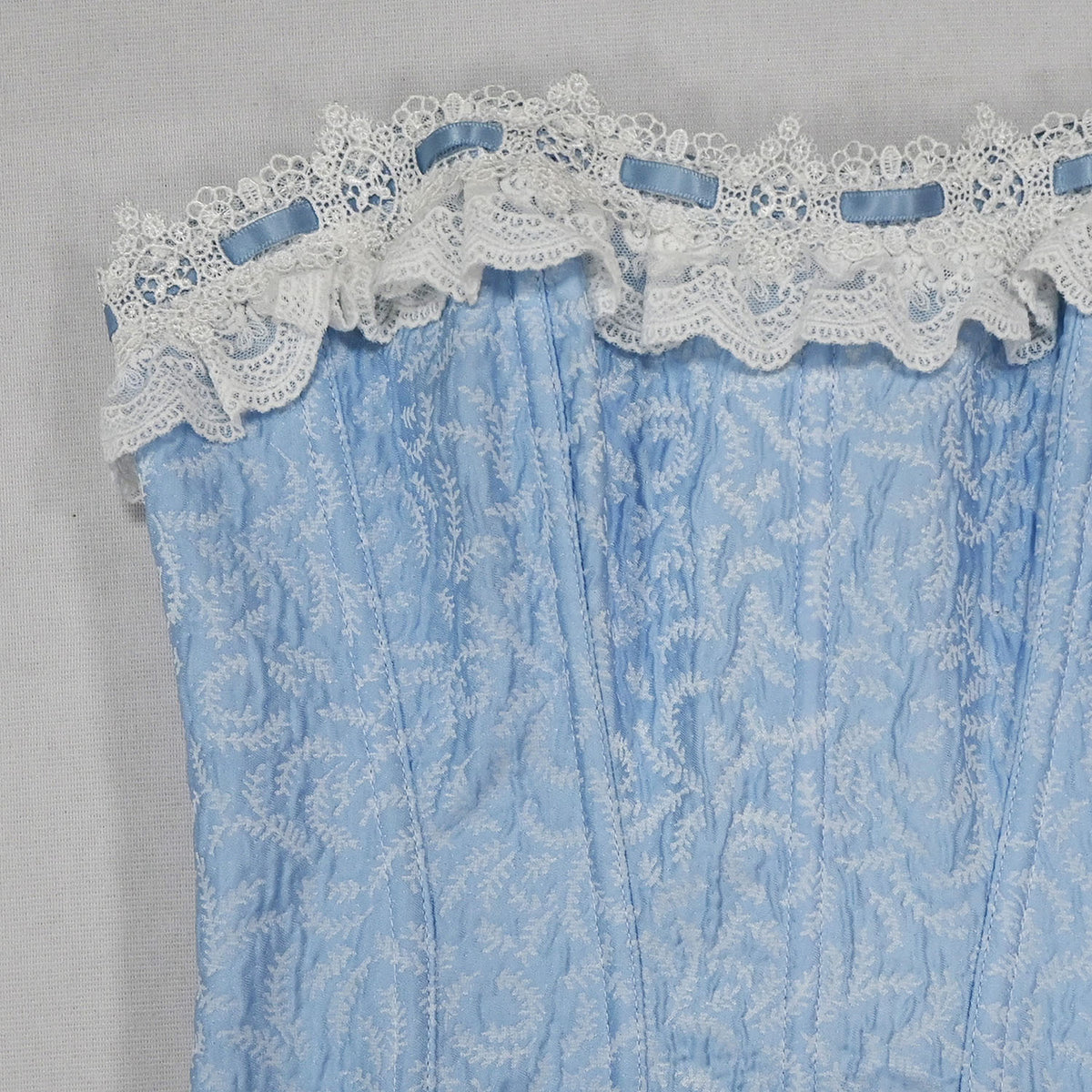 A close-up of a light blue fabric panel with white floral embroidery and a white lace trim with small blue ribbon accents, laid flat against a light gray background.