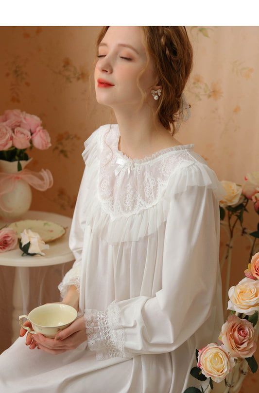A woman wearing a white long-sleeve nightgown with delicate lace trim and ruffled details, holding a teacup in a softly decorated room with pink roses.