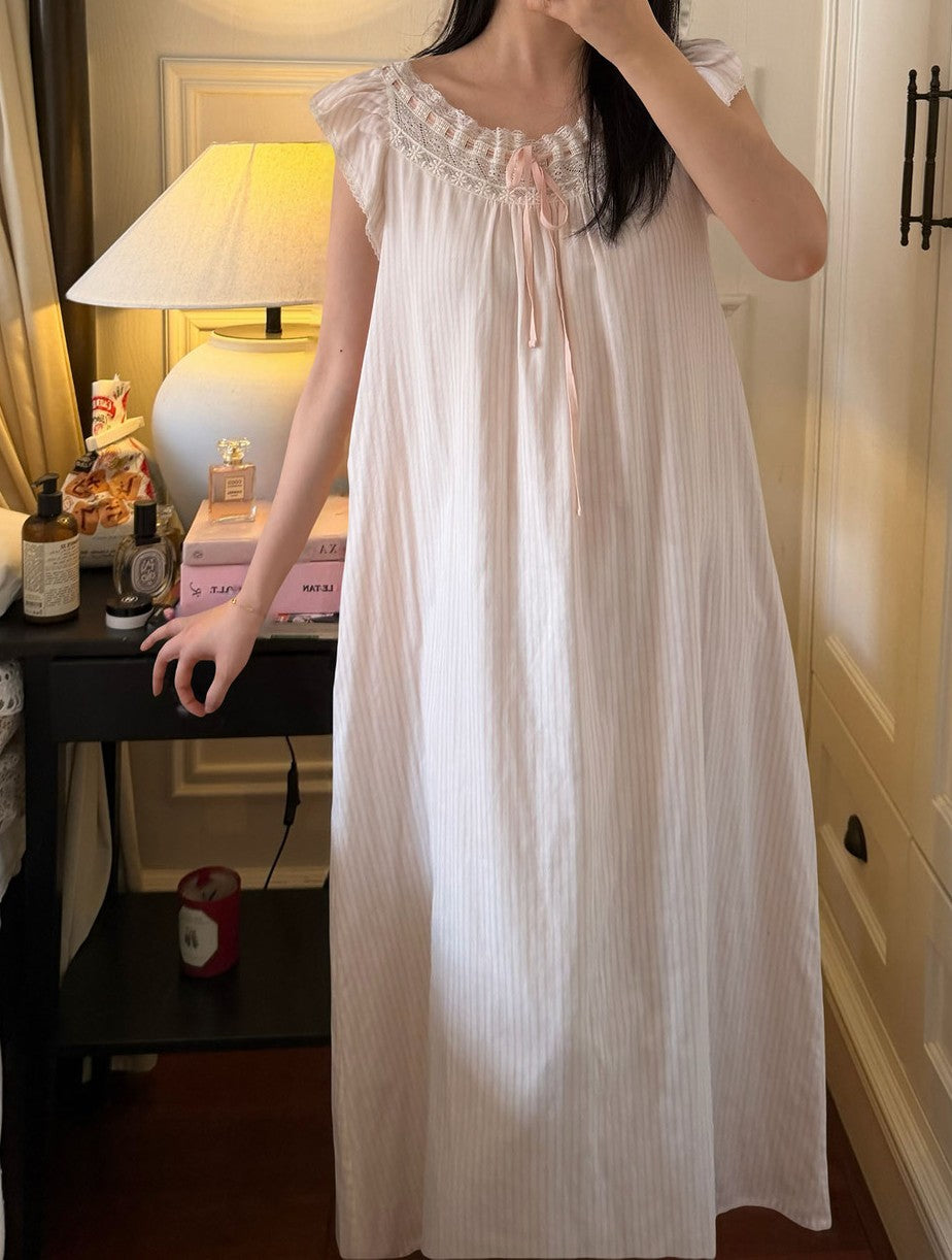 A woman wearing a long white ribbed nightgown with a ruffled lace-trimmed neckline and a small bow at the center front, standing indoors next to a bedside table with a lit lamp.