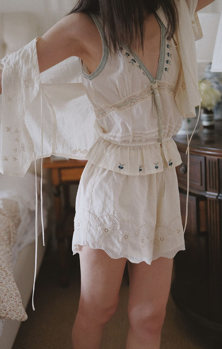 A woman wearing a cream-colored two-piece matching set consisting of a sleeveless top with lace trim and a matching ruffled mini skirt, styled with a matching sheer long-sleeve shirt in a cozy bedroom setting.
