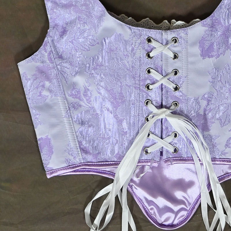 A close-up of a lavender and white jacquard corset top with lace-up front, silver metal eyelets, and white satin ties, laid flat on a brown surface.