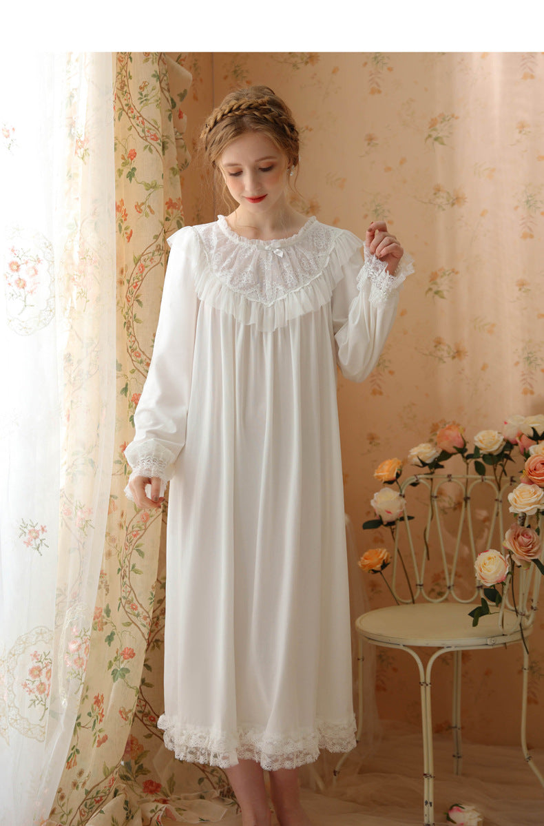 A woman wearing a long white nightgown with a ruffled lace neckline and cuffs, standing in a vintage-style room with floral wallpaper, sheer curtains, and a small table with flowers.