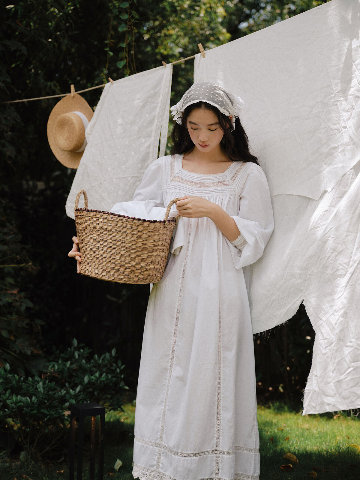 A woman in a long white dress and lace head covering stands outdoors holding a woven basket, with white laundry hanging on a line behind her in a garden setting.