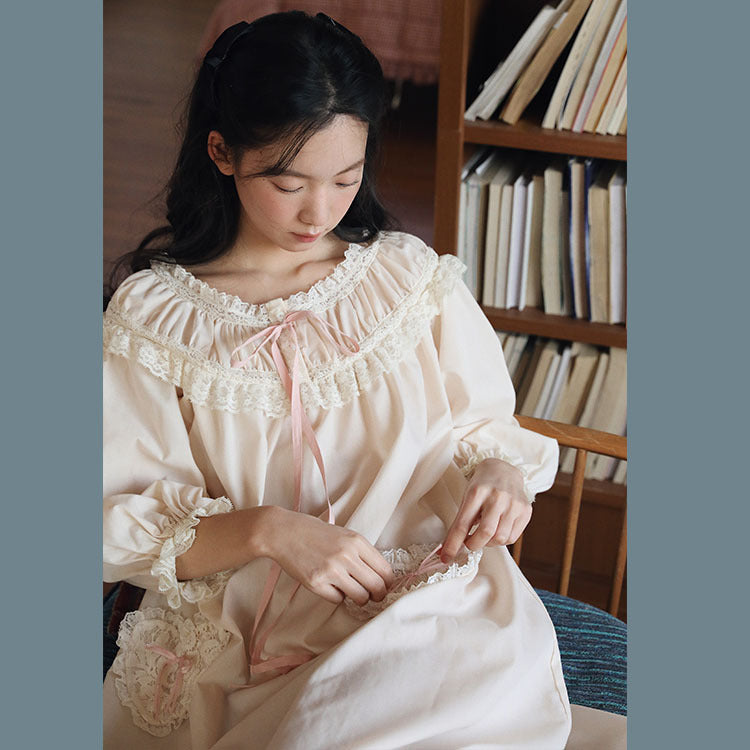 A young woman with long black hair sits in a cozy room, wearing a cream-colored long-sleeve nightgown with intricate lace trim and a pink ribbon tie. She is looking down while adjusting the fabric of the nightgown.