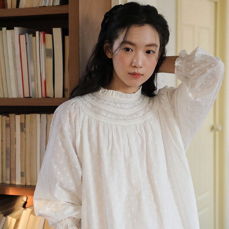 A woman wearing a white long-sleeve blouse with a ruffled mock neck and subtle polka dot pattern, standing in front of a wooden bookshelf.