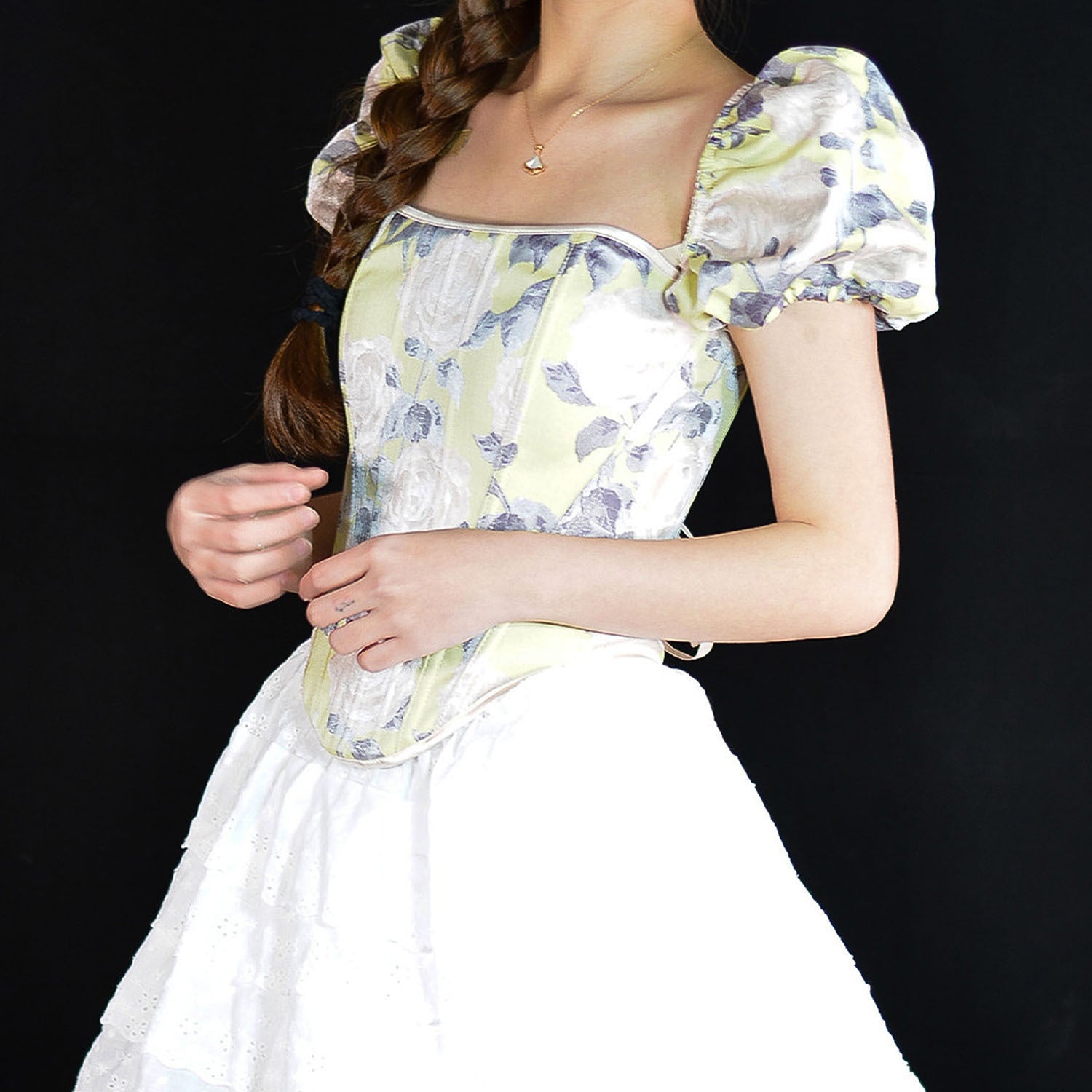 A woman wearing a vintage-style corset dress with a pale yellow and lavender floral corset top, puffed sleeves, and a white skirt, set against a solid black background.