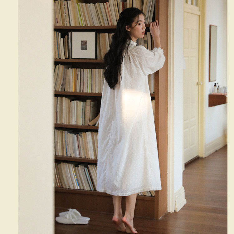 A woman stands barefoot in a warmly lit room with wooden floors and a bookshelf, wearing a loose white long-sleeve midi dress. White slippers are placed on the floor nearby.