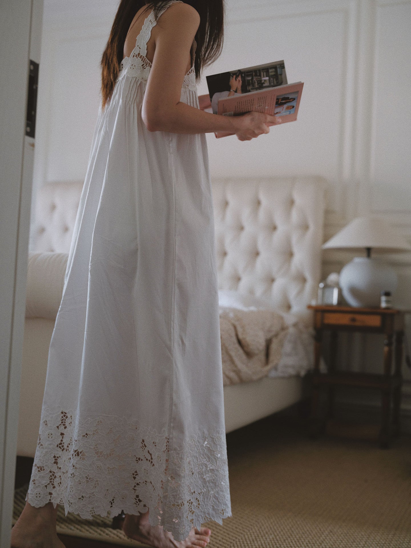 A woman in a white sleeveless nightgown with lace trim stands in a bedroom holding a magazine. The room features a tufted cream headboard, a bedside table with a lamp, and a beige carpet.
