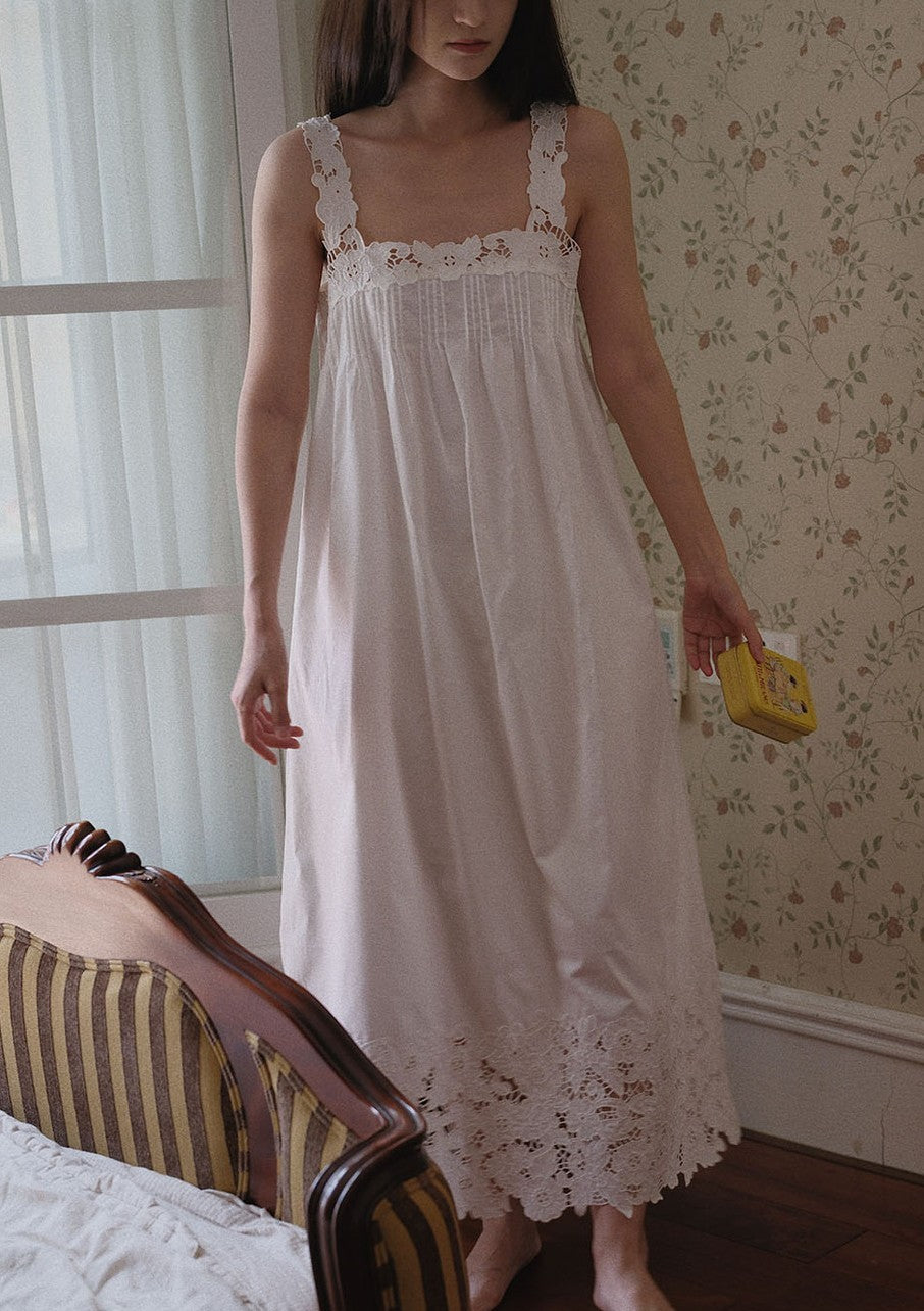 A woman wearing a long white nightgown with lace trim and embroidered details, standing in a bedroom with floral wallpaper and a striped armchair.