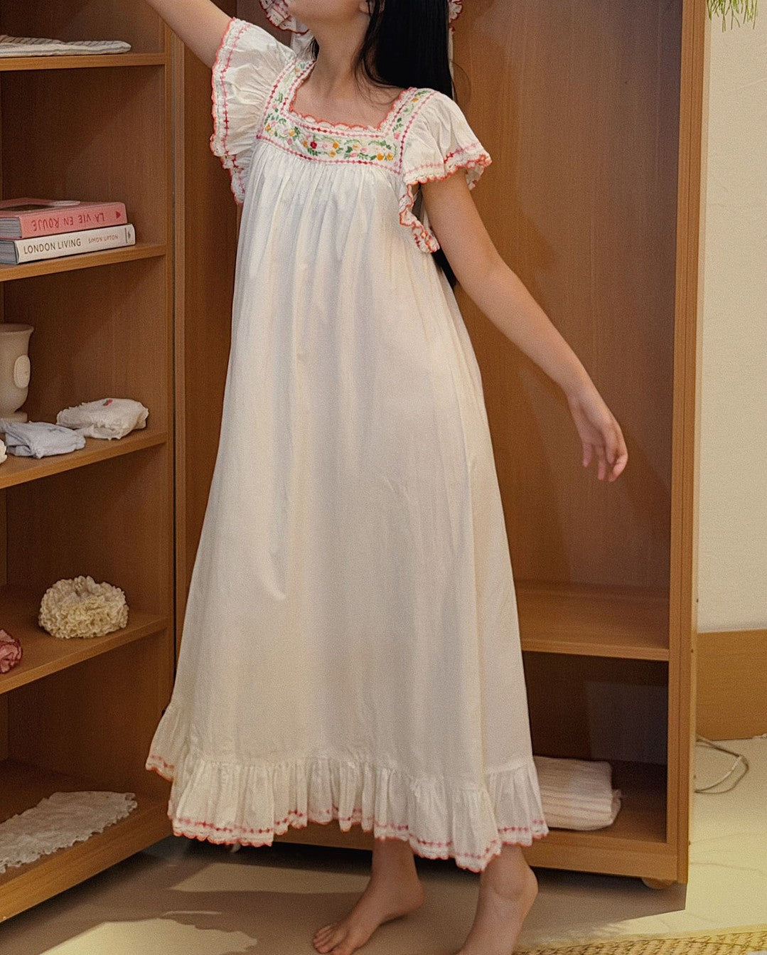 A woman wearing a long white nightgown with pink and multicolor floral embroidery on the bodice and ruffled trim, standing in front of a wooden shelving unit.