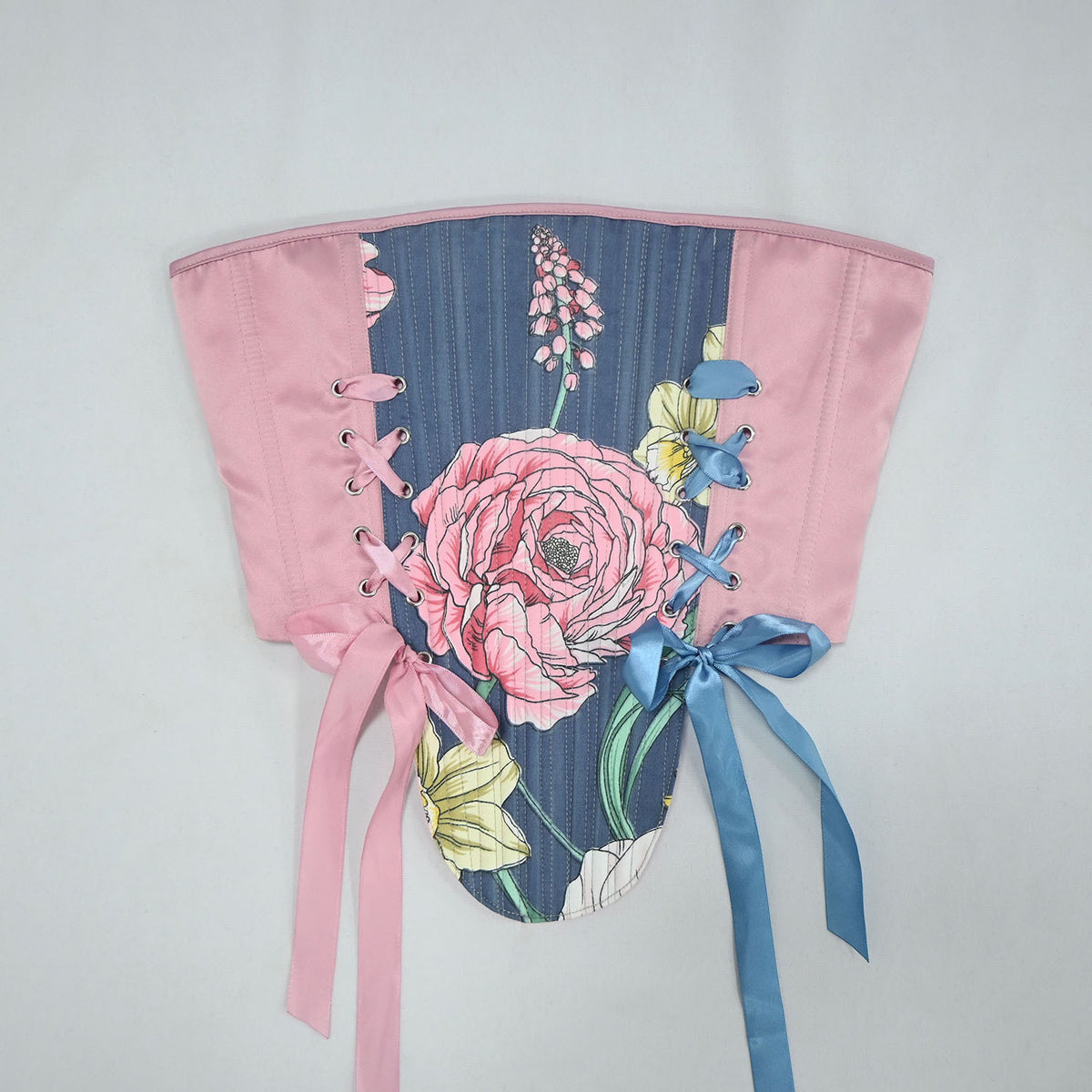 A floral corset top with a pink satin waistband and flared sleeves, featuring a blue floral print with a large pink rose, yellow flowers, and silver lace-up eyelets, finished with pink and blue ribbons.