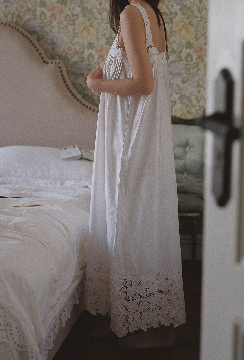 A woman wearing a long white nightgown with lace trim, standing in a bedroom next to a bed with white bedding.