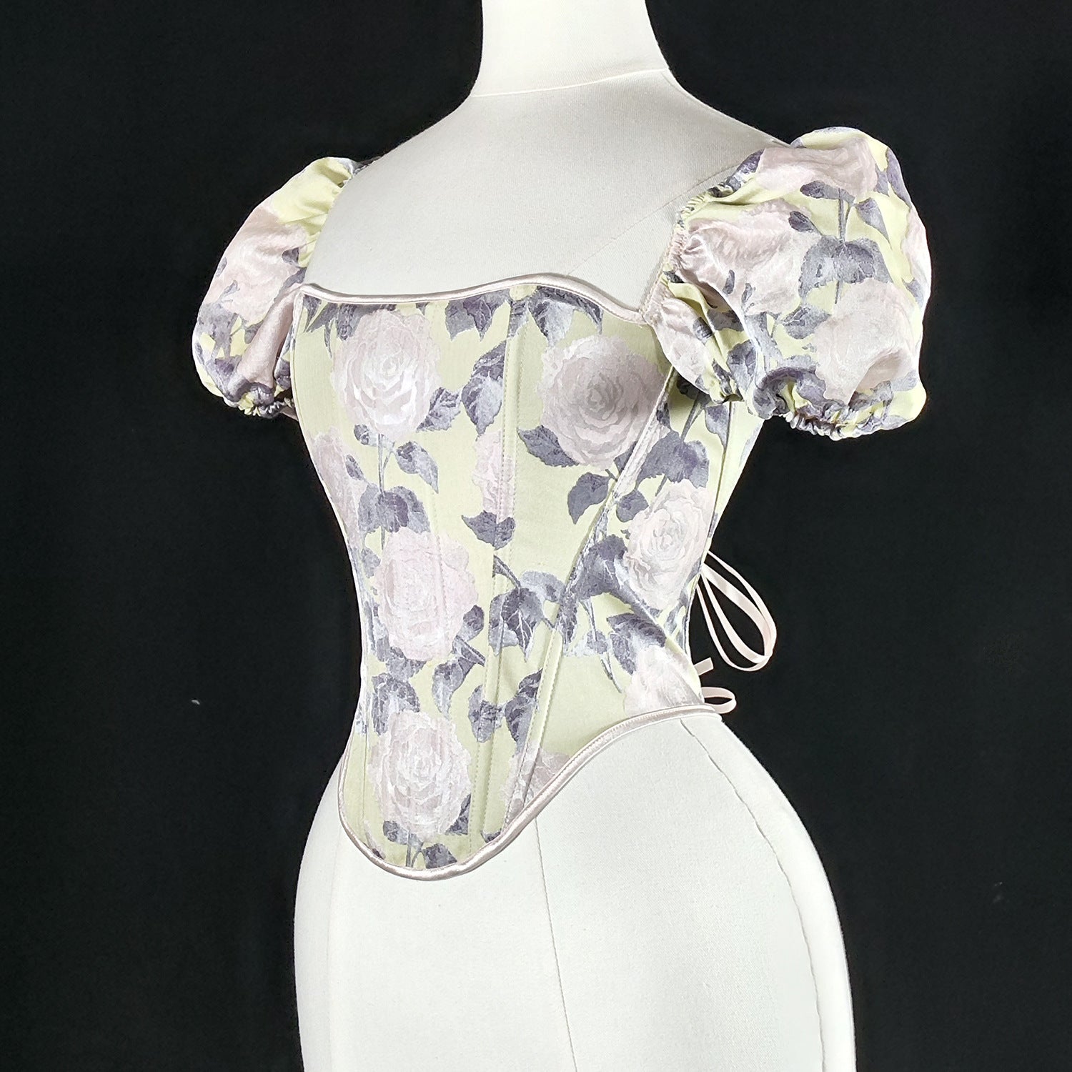 A structured corset-style top with puff sleeves, displayed on a white mannequin torso against a black background. The fabric features a pale sage green base with a floral rose pattern in muted lavender-gray and soft pink tones.