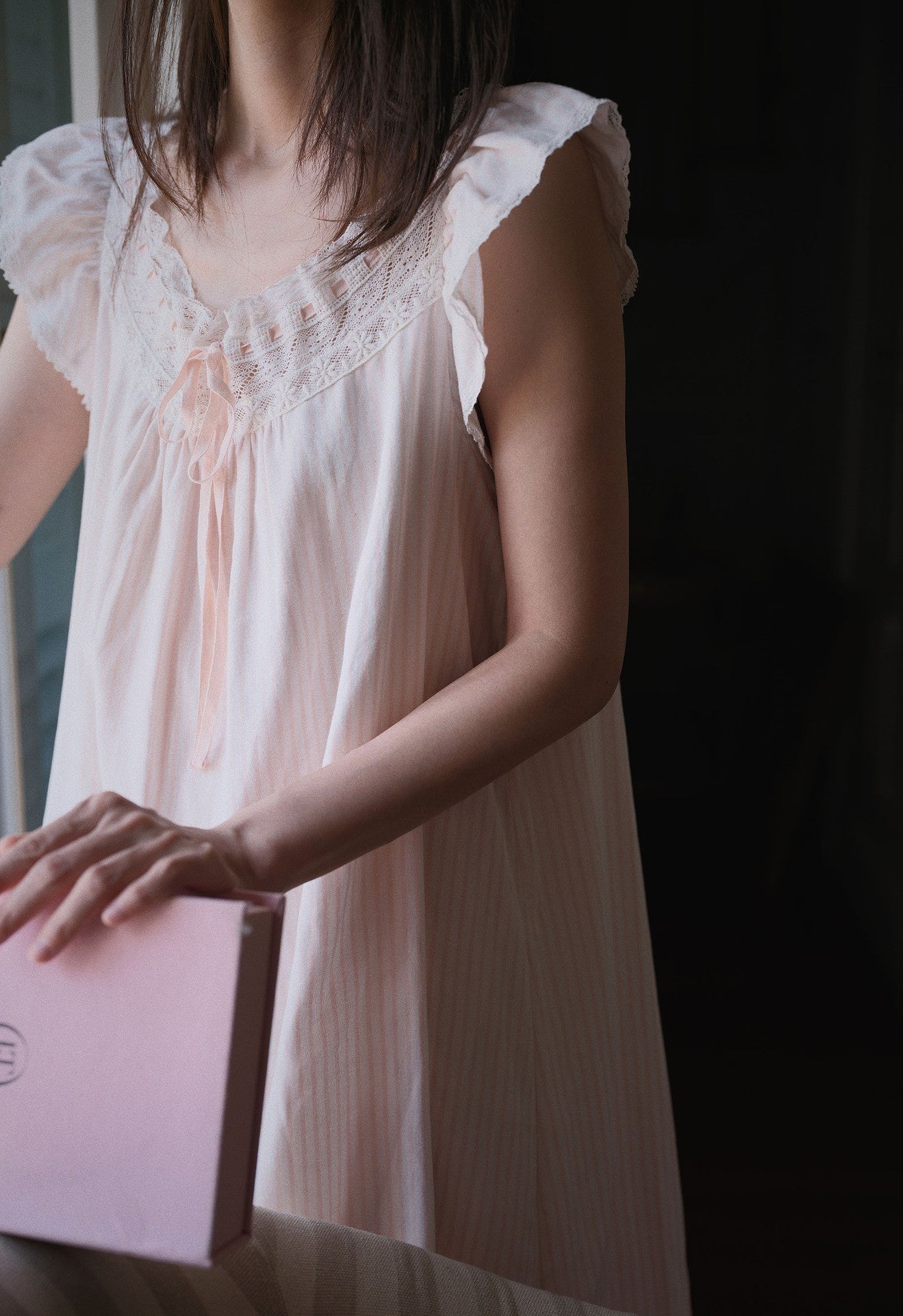 A woman wearing a pale blush pink sleeveless nightgown with lace trim and a tie at the neckline, holding a matching pink box in a dimly lit setting.