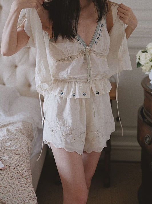 A woman wearing a cream-colored two-piece lingerie set with lace and embroidery, standing in a bedroom setting.