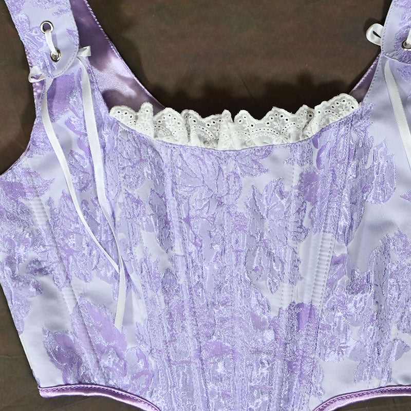 A lavender and white floral jacquard corset bustier top with white lace trim and white shoulder ties, laid flat on a dark brown surface.