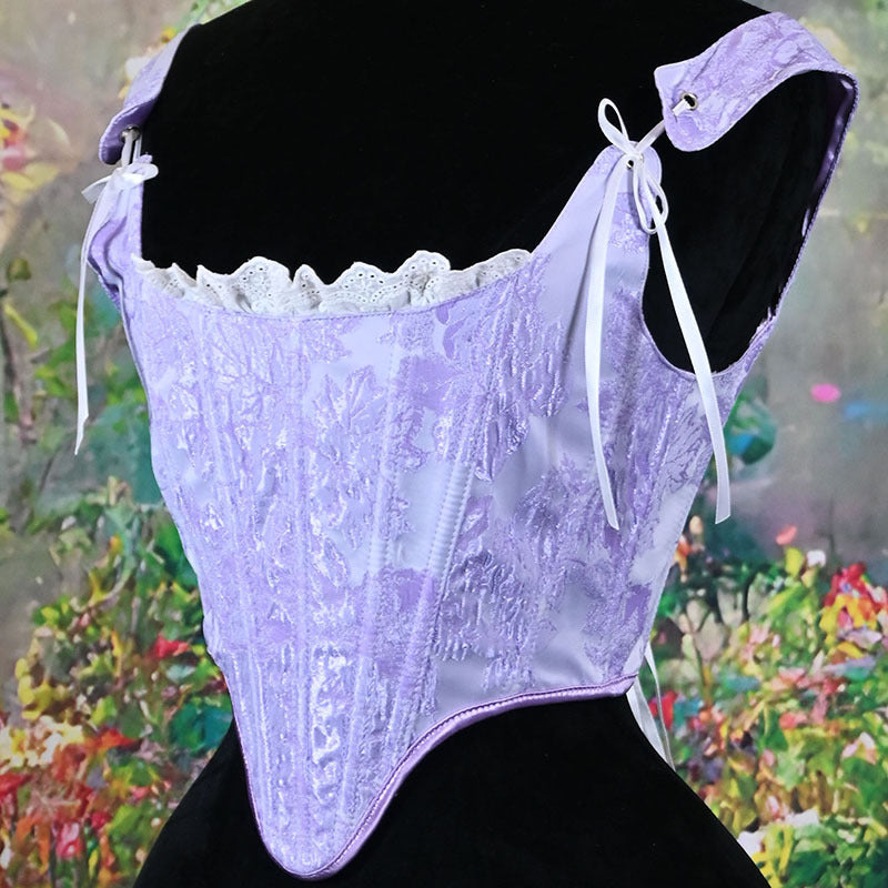 A lavender lace and satin corset top displayed on a black mannequin torso, featuring floral embroidery, white lace trim, and white tie straps, set against a colorful painted garden background.