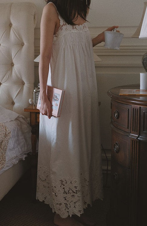 A woman wearing a long white lace-trim nightgown or smocked dress stands in a bedroom holding a mug and a book.