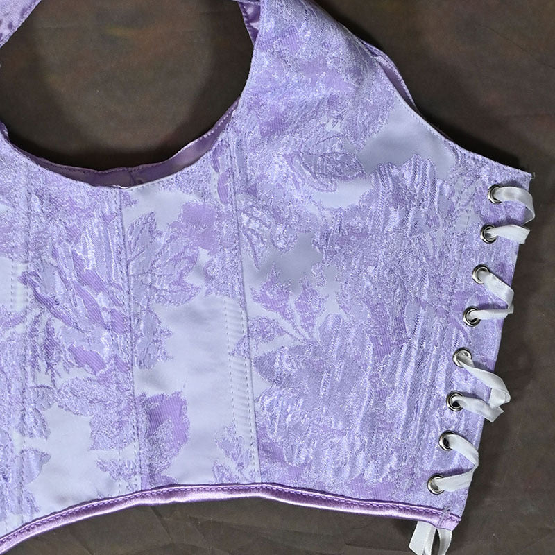 A close-up of a lavender and white patterned corset bustier with white lace-up ties on the side, laid flat on a dark brown surface.
