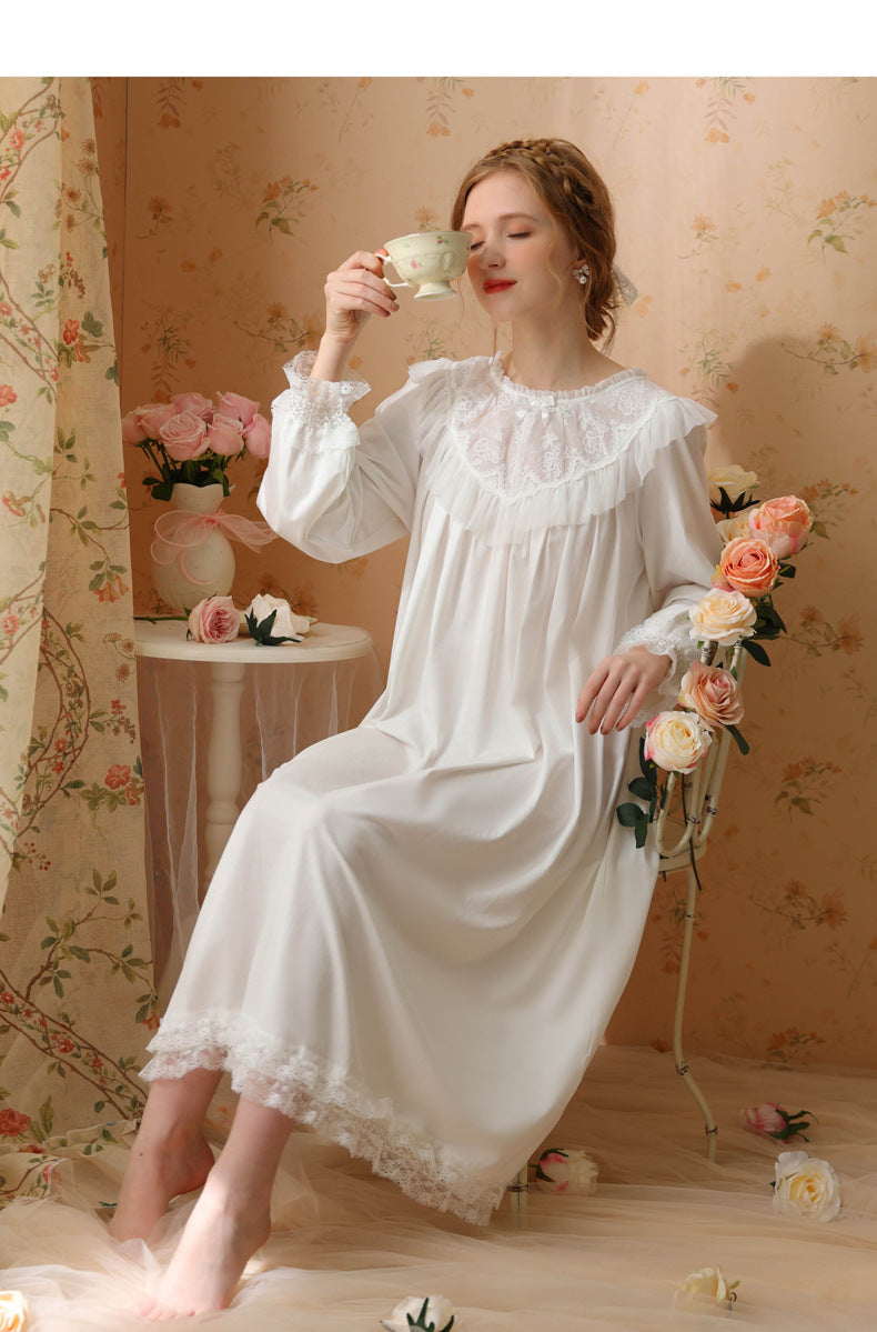 A woman wearing a long white satin nightgown with lace trim sits on a chair in a vintage-style room, holding a teacup. The background features floral wallpaper, a small table with flowers, and a curtain.