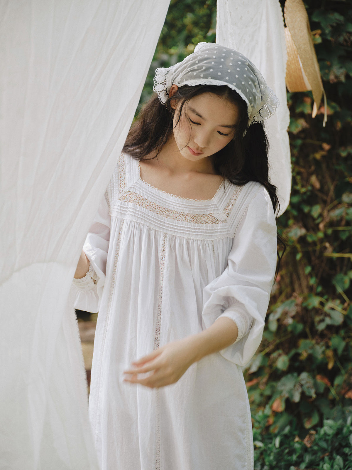 A woman wearing a white long-sleeve embroidered nightgown and a white polka-dot headscarf, standing in front of a white hanging curtain with green foliage in the background.